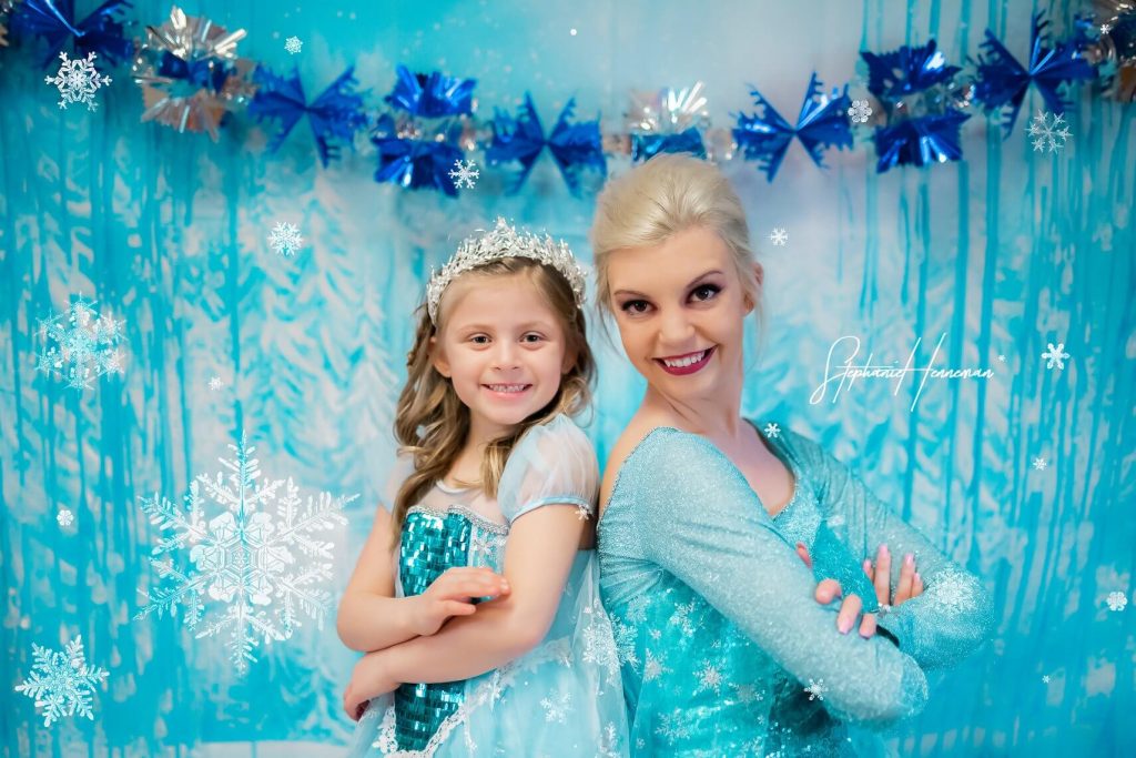 Queen Elsa and a young girl pose for a picture against a backdrop at a princess party