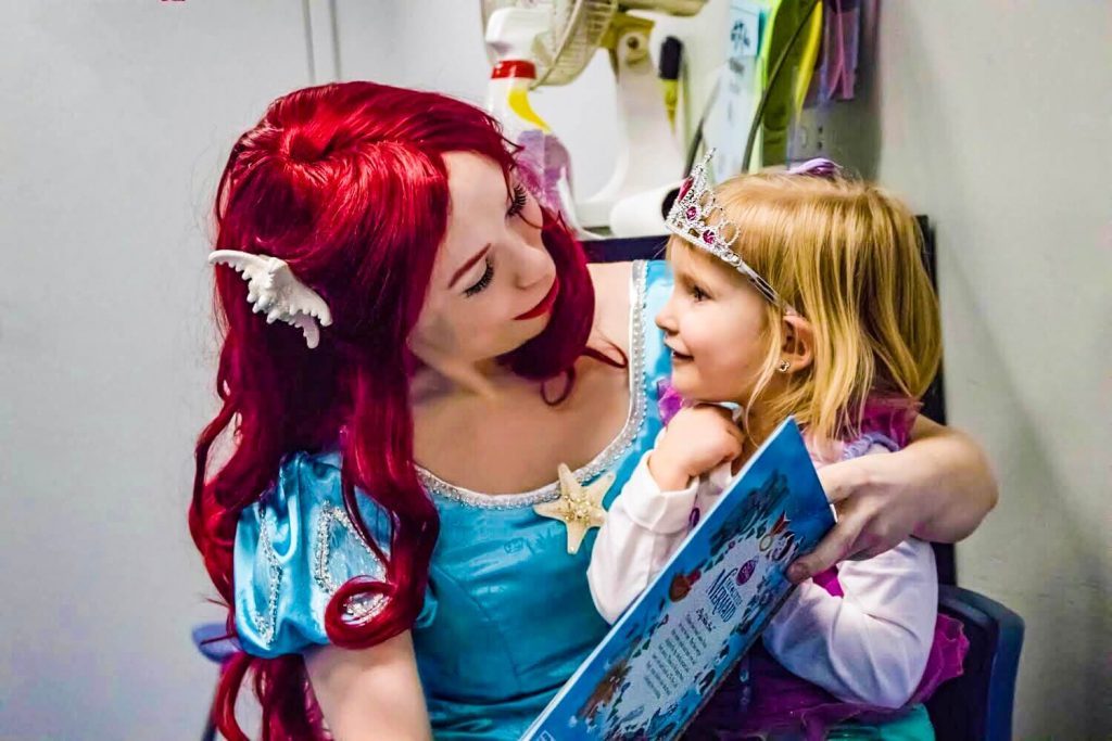 Princess Ariel holds a little girl as they read a story together at a Little Mermaid birthday party