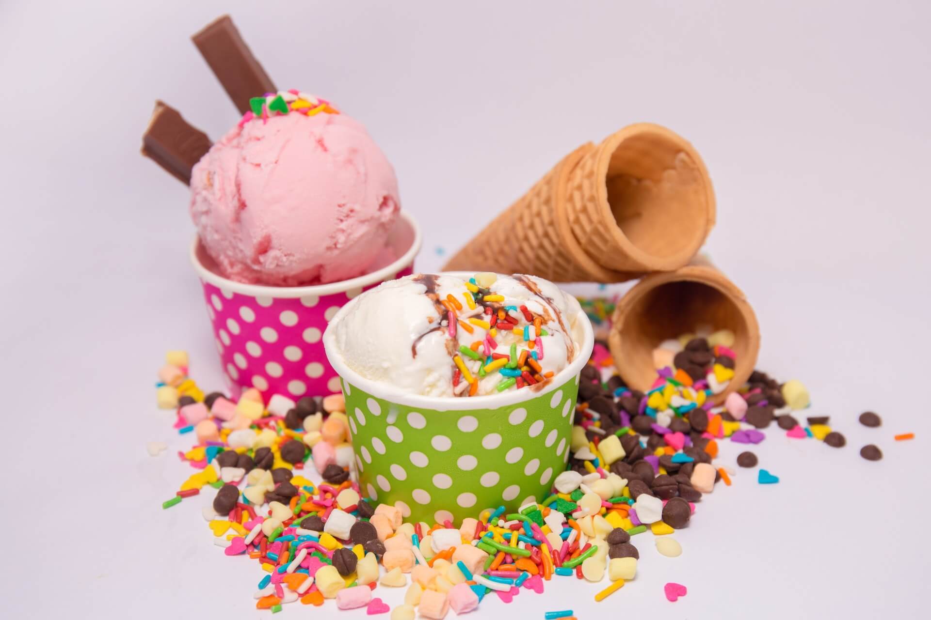 This picture features two cups of ice cream with several ice cream cones in the background. These treats are surrounded by sprinkles, chocolate chips, and other ice cream toppings.